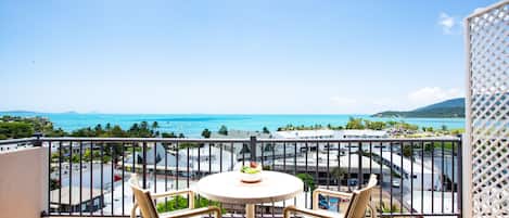 A balcony with a table and two chairs, overlooking a coastal view with buildings and a road.