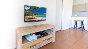 Uma sala de estar com um armário de entretenimento de madeira, uma TV de tela plana exibindo uma cena de praia, e uma área de cozinha com armários claros e um banquinho de madeira.