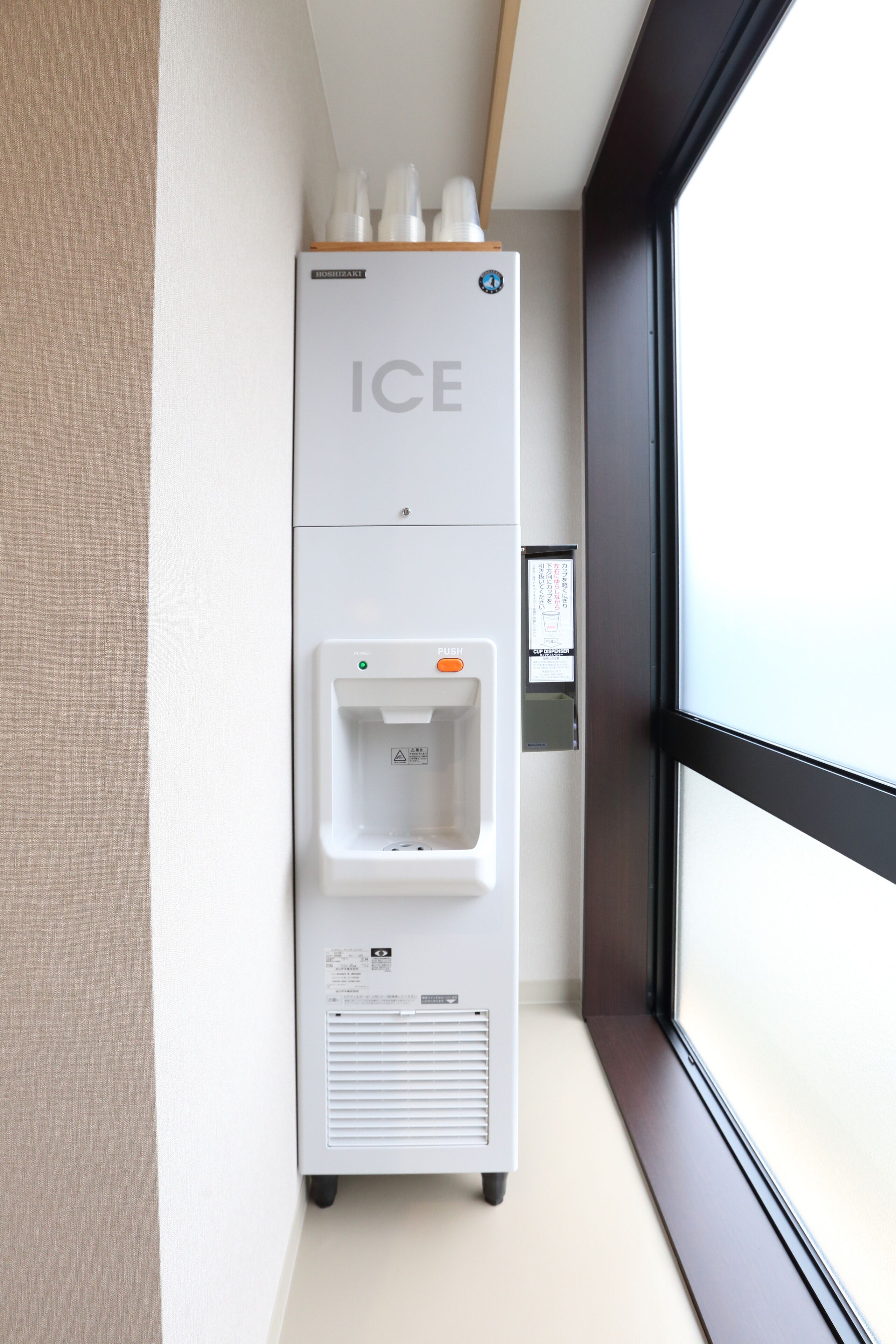 Een witte, aan de muur bevestigde ijsdispenser met bedieningspaneel en een waterdispenser, geplaatst in een hoek met een raam.