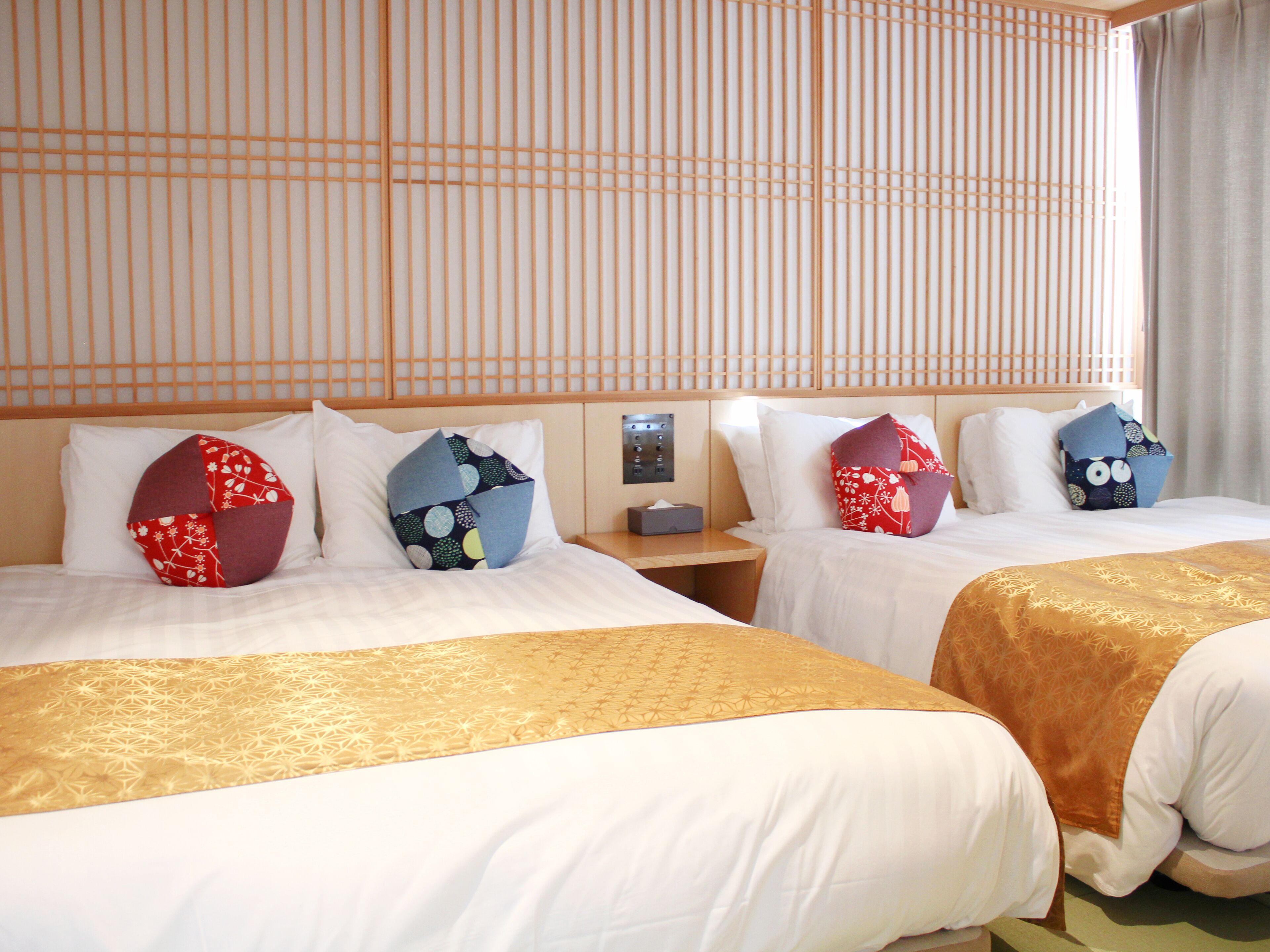 Une chambre d’hôtel avec deux lits, chacun agrémenté d’un coussin à motifs distincts, une table de chevet avec une lampe et une vue sur l’extérieur par une fenêtre munie de rideaux.