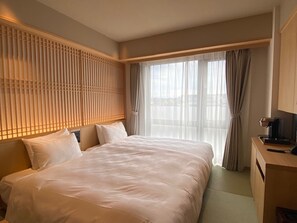 Ein Hotelzimmer mit einem großen Bett, einem Schreibtisch mit Lampe und Blick auf die Stadt durch große Fenster.