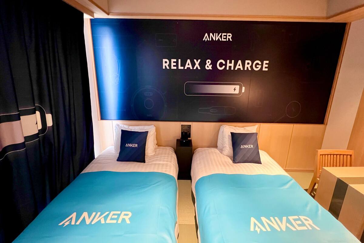 A hotel room with two beds, each with a blue bedspread and pillows labeled "ANKER". A large screen on the wall displays "ANKER RELAX & CHARGE".
