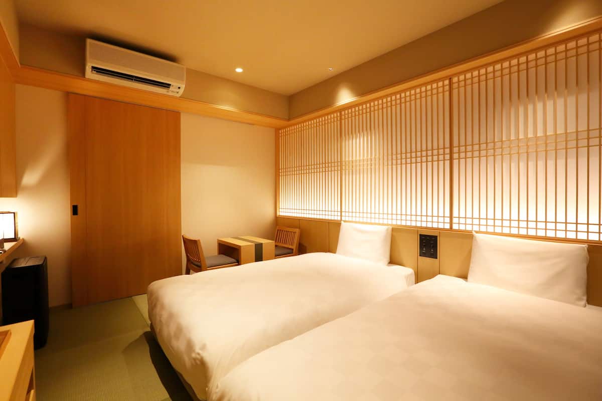 A hotel room with two beds, a wooden wardrobe, a small table, a chair, and a traditional Japanese shoji screen.