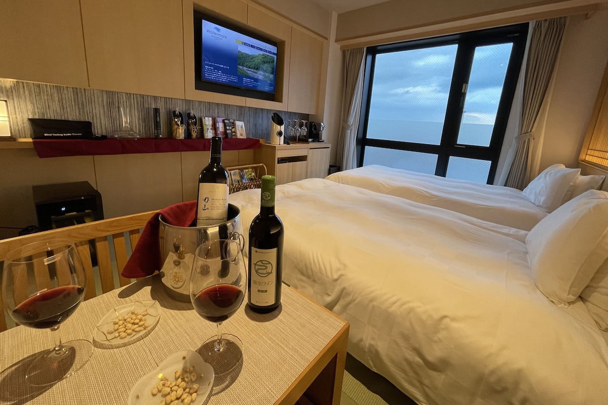A hotel room with a bed, a TV mounted on the wall, a table with wine glasses and bottles, and a view of the sea through the window.