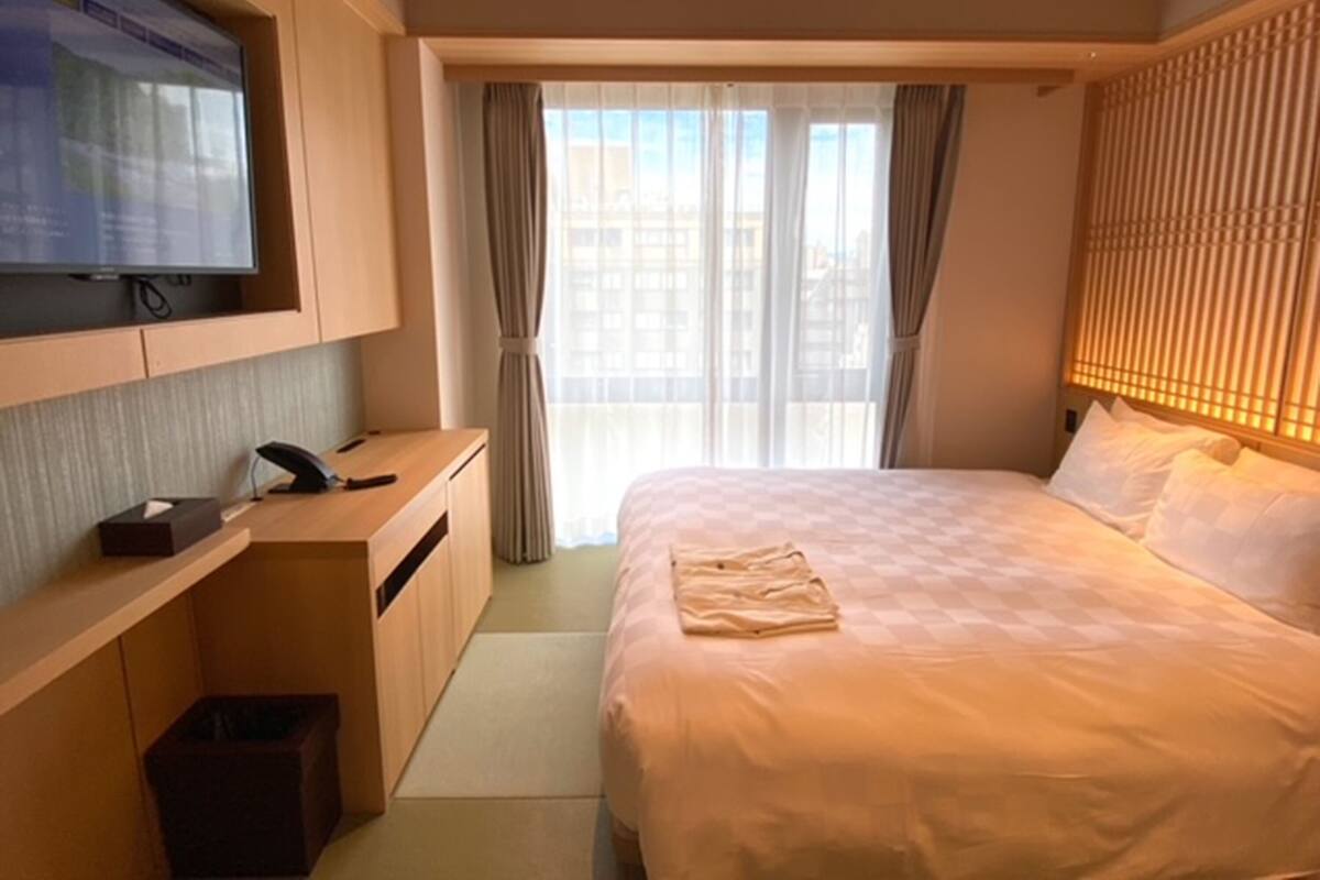 A hotel room with a bed, a desk with a television, and a view of buildings through the window.