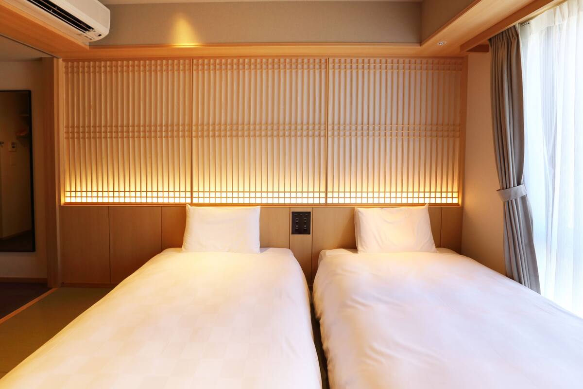 A hotel room with two beds, a wooden headboard with a light panel, and a window with curtains.