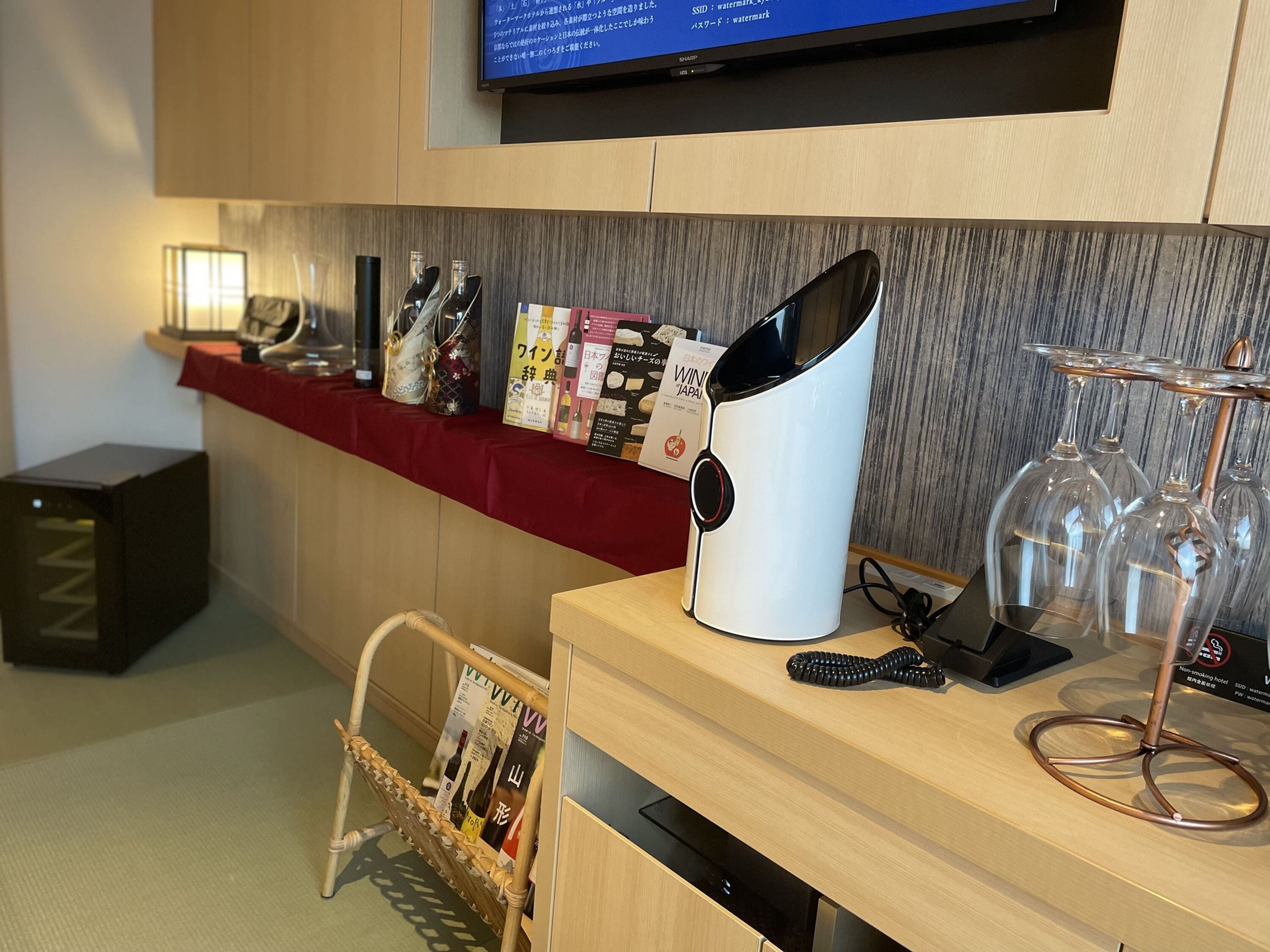 A hotel reception area with a wooden counter, a television mounted above, magazines and brochures on the counter, a wine cooler, and a glass display case with wine glasses.