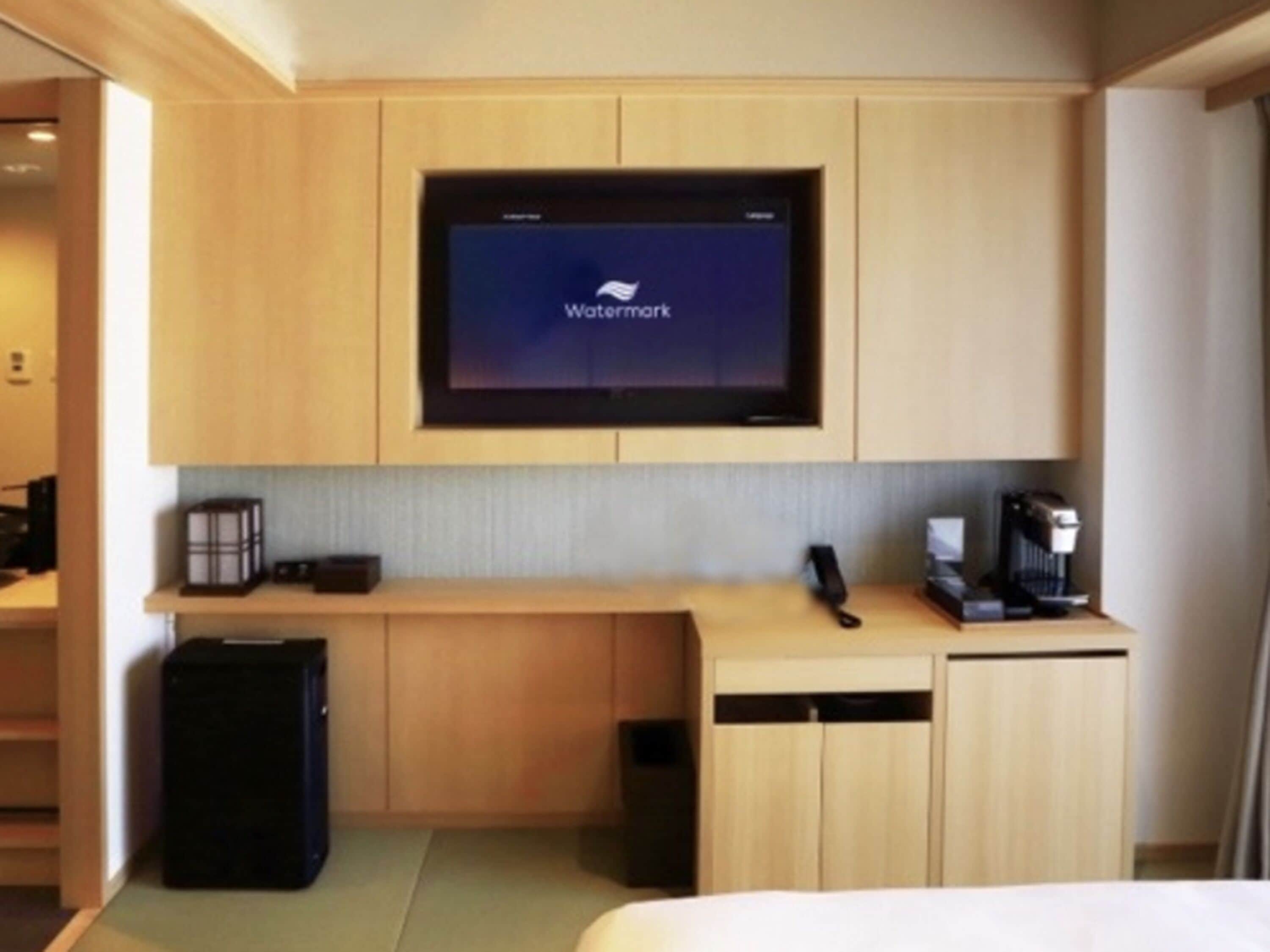 Habitación de hotel con cama, escritorio con monitor que muestra 'Watermark', cafetera y maleta.
