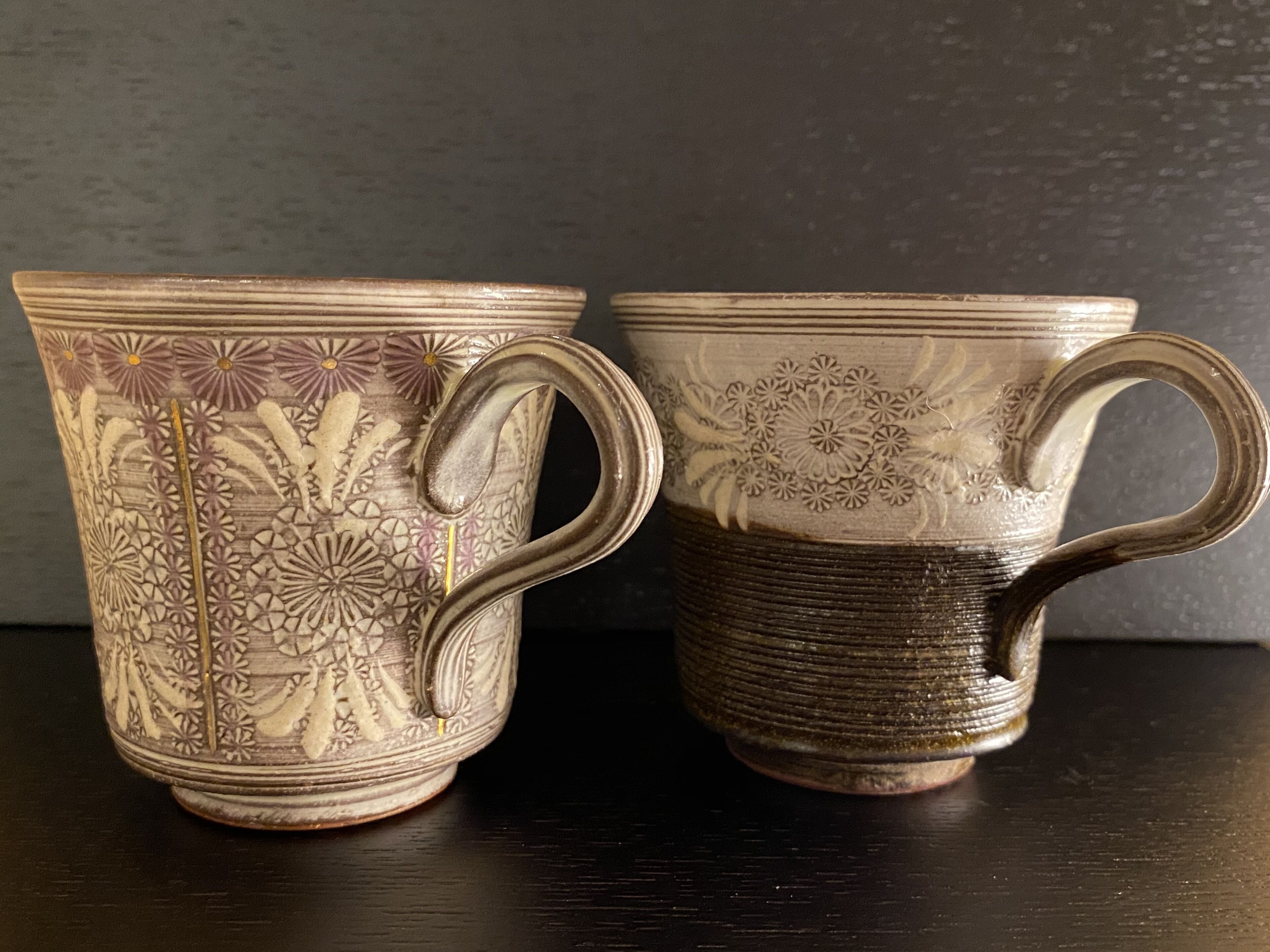 Deux tasses en céramique ornées de motifs floraux complexes et dotées d’anses, posées sur une surface sombre.