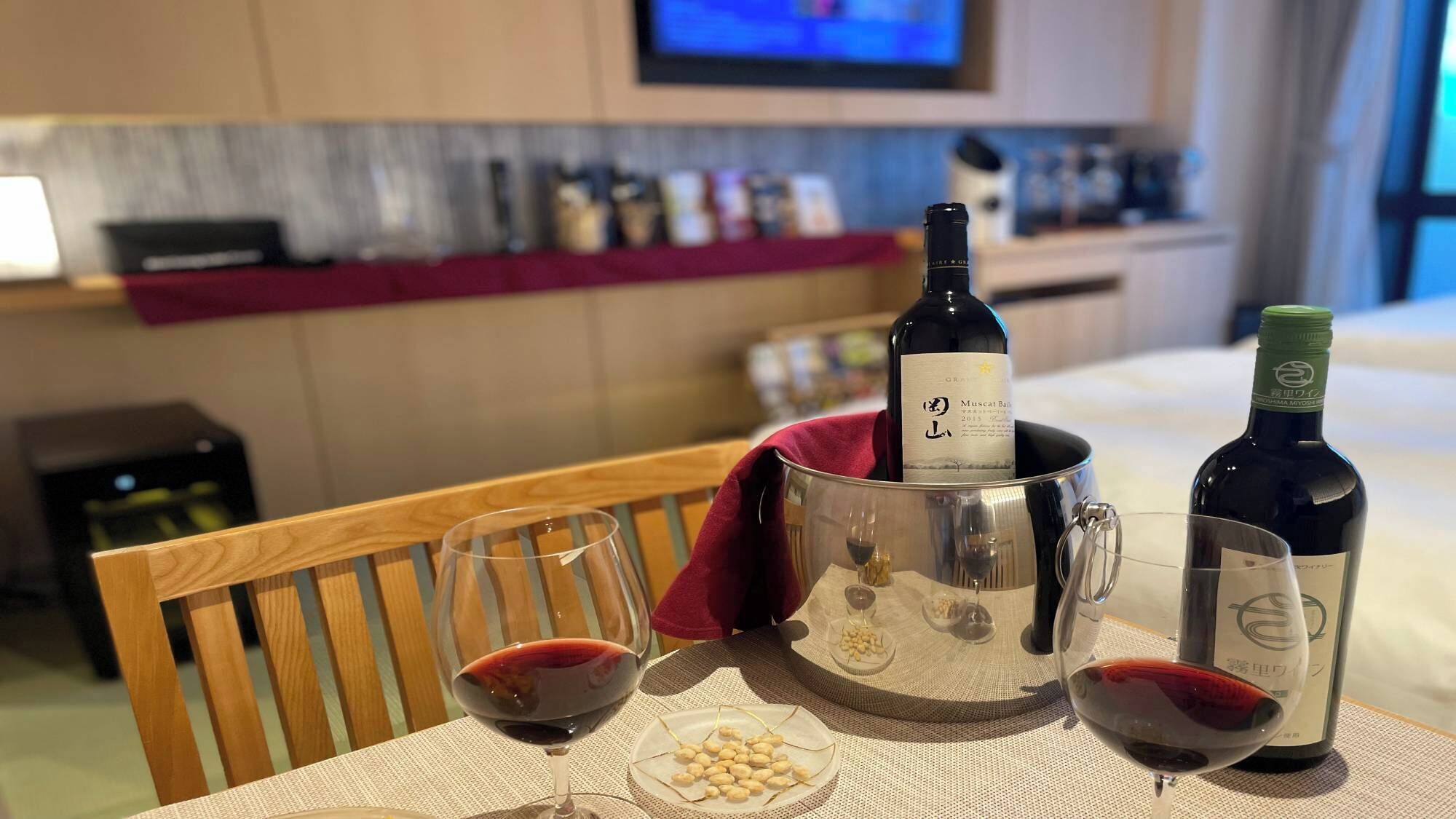 Une chambre d’hôtel avec une table à manger dressée, comprenant des verres à vin, une bouteille de vin et une assiette de cacahuètes.