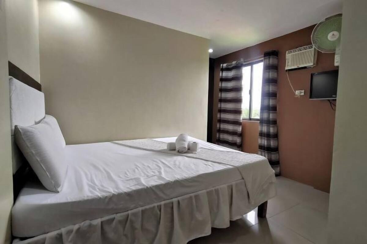 A neatly made bed with pillows, a bedside table, a wall-mounted TV, and a standing fan.