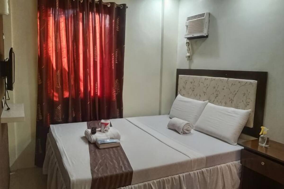 A hotel room with a bed, a nightstand, a wall-mounted air conditioner, and red curtains.