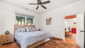 Une chambre à coucher comprenant un lit, une tête de lit en bois, une table de chevet, un ventilateur de plafond, une fenêtre et une porte donnant sur une autre pièce.