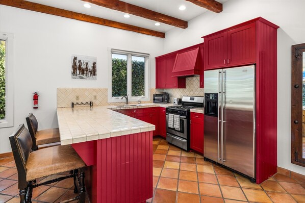 A kitchen with red cabinets, a stainless steel refrigerator, a stove, and a window with a view of greenery.