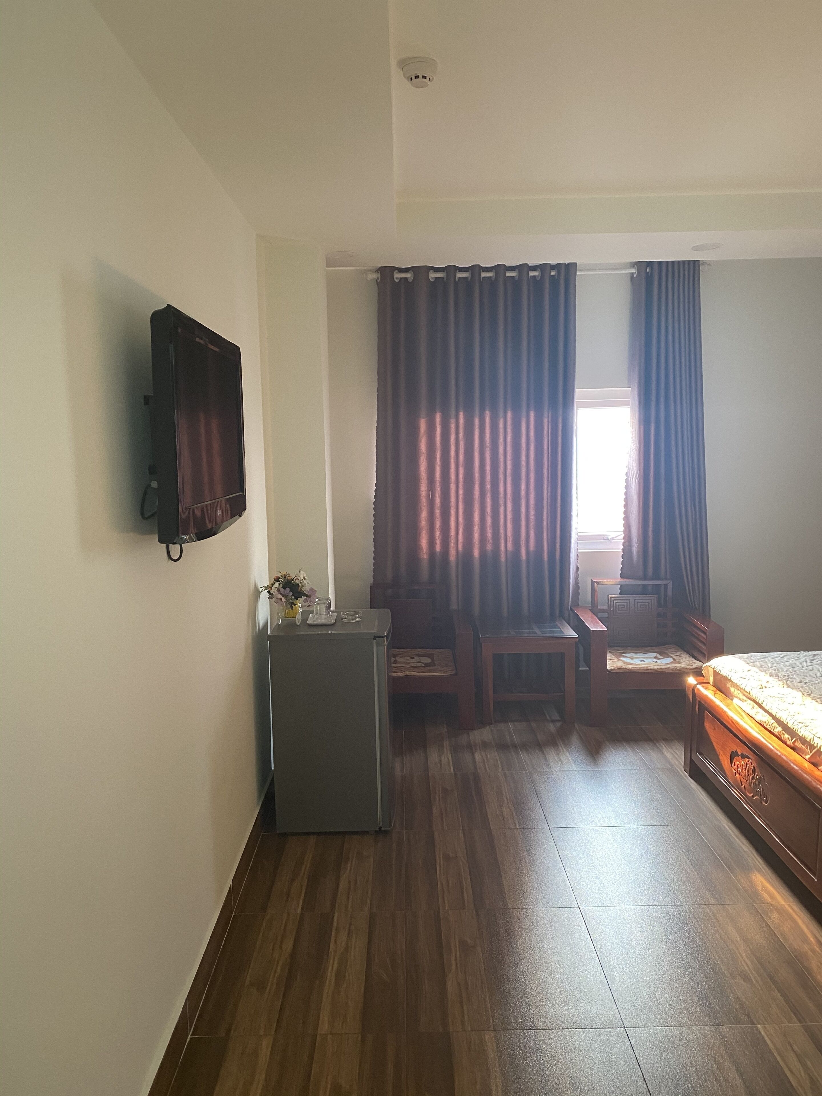 A small, well-lit room with a bed, a desk, a chair, and a television mounted on the wall.
