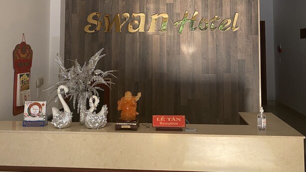 A reception desk with a wooden backdrop, a sign reading "Swan Hotel," and various decorative items including a plant, a statue, and a sign for the reception. - Swan Hotel (Da Nang)