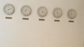 Un mur sur lequel sont accrochées cinq horloges, chacune portant le nom d’une ville : Paris, Tokyo, Hanoï, Londres et New York. En dessous des horloges, on trouve un banc en bois doté d’un dossier.