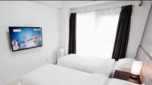 A hotel room with two beds, a TV mounted on the wall, a bedside table with a lamp, and a window with curtains. - Hostel 758 Nagoya2B (Nagoya)