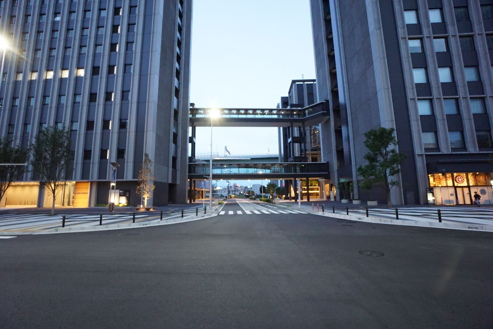 A modern urban street with tall buildings, a pedestrian crossing, and a bridge overhead.