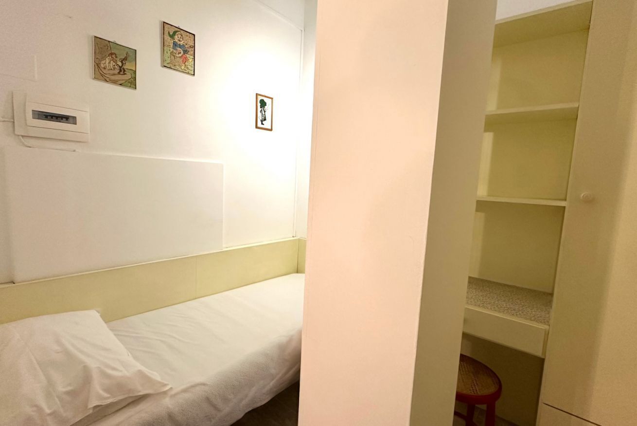 A single bed with a white mattress and pillow, a wall-mounted heater, and a small stool.