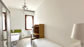 Una stanza piccola e ben illuminata con un letto, una scrivania e una sedia. C'è un termosifone sotto la finestra e un lampadario appeso al soffitto.