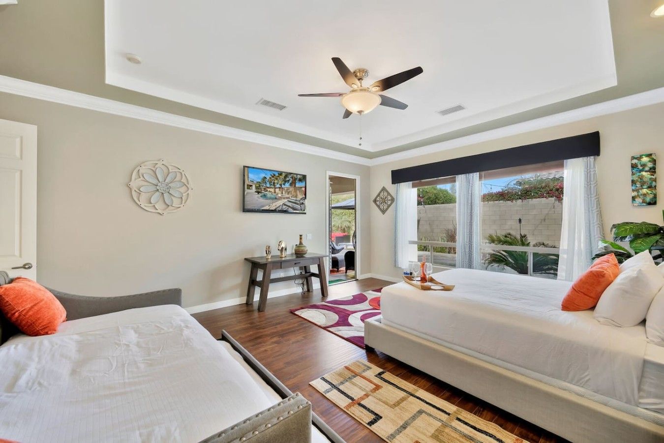 A hotel room with two beds, a ceiling fan, a painting, a desk, and a view of the outdoors.