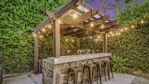 An outdoor bar area with a stone countertop, bar stools, and a pergola with string lights.