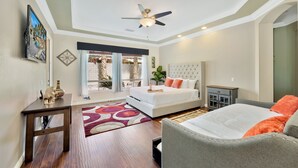 A bedroom with a bed, a gray armchair, a nightstand, a ceiling fan, a window with curtains, and a view of the outdoors.