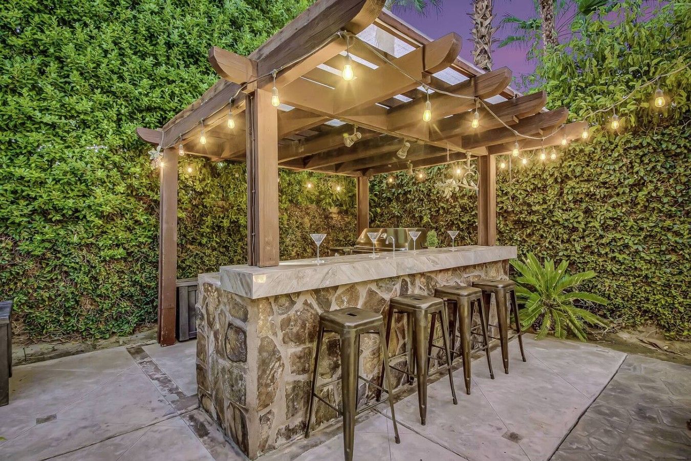 An outdoor bar area with a stone counter, bar stools, and a pergola with string lights.