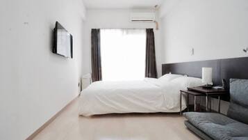 A hotel room with a bed, a bedside table with a lamp, a sofa, and an air conditioning unit.