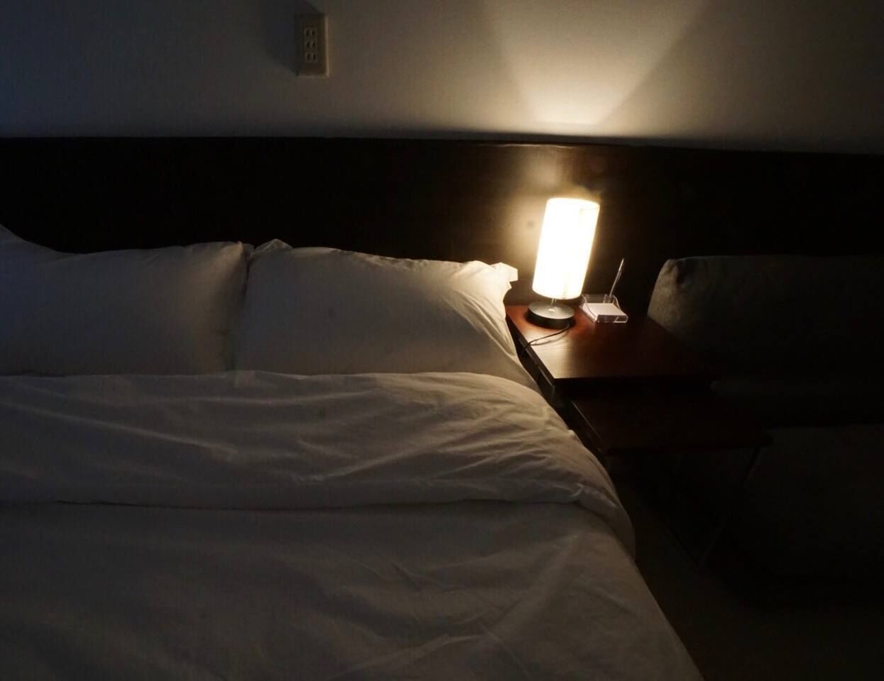A dimly lit hotel room with a single bedside table, a lamp, and a bed with white linens.