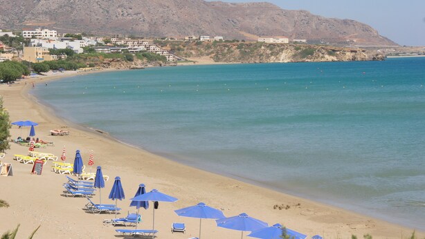 A beach with rows of sunbeds and umbrellas, a clear blue sea, and buildings in the background. - Makrigialos: big family apartment, 2 bedrooms, sea views, WiFi (Makrigialos)