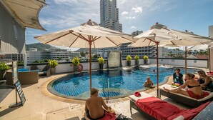 Un área de piscina en la azotea con sillas de descanso y sombrillas, rodeada de plantas en macetas y vistas panorámicas de la ciudad.