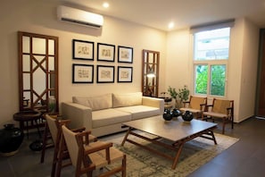 A living room with a beige sofa, wooden chairs, a coffee table, and framed artwork on the wall.