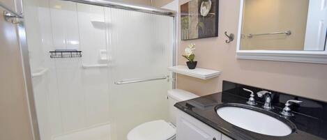 A bathroom with a glass shower enclosure, a white toilet, a black sink with a mirror above it, and a small shelf with a flower.