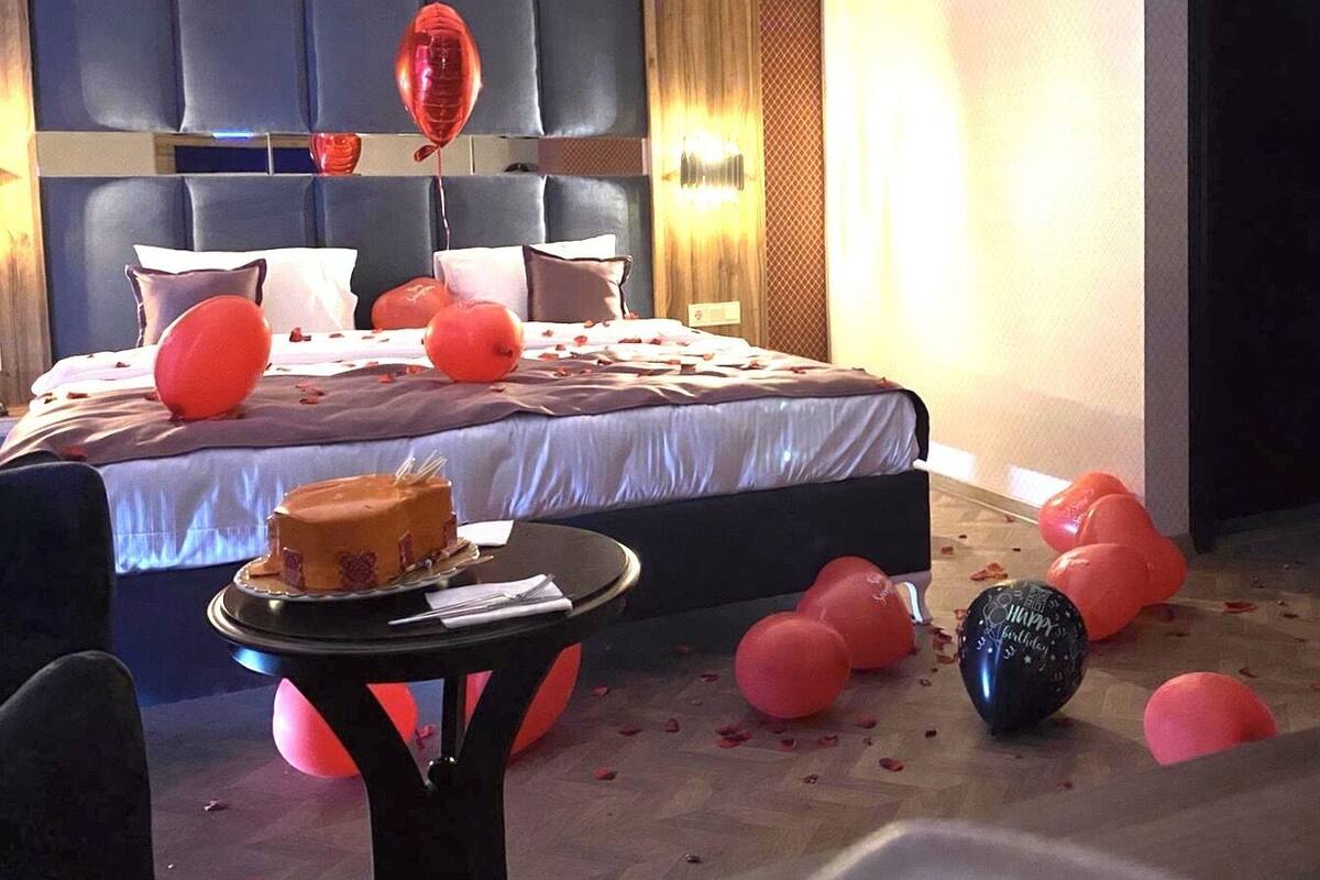 A hotel room with a large bed, a small round table with a cake, and scattered balloons.