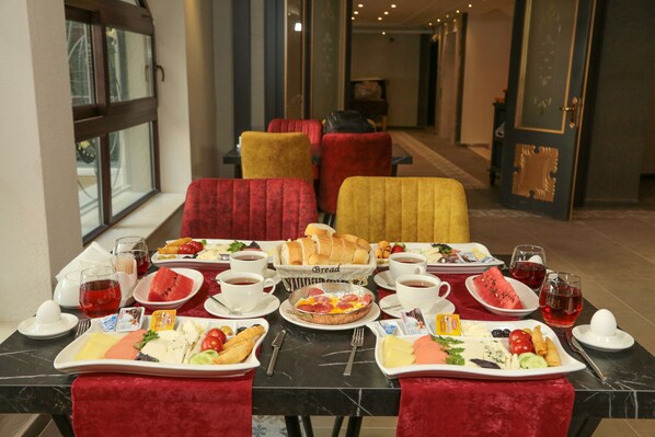 Un desayuno con una variedad de platillos, como pan, frutas y pastelería, dispuestos sobre una mesa de mármol con sillas tapizadas en rojo.