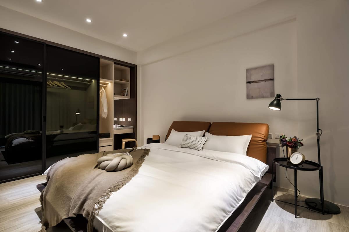 A modern bedroom with a large bed, a wardrobe, a bedside table with a lamp, and a wall-mounted clock.