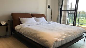 A modern bedroom with a large bed, a bedside table, a lamp, and a view of greenery outside.