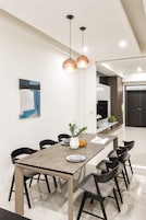Uma área de jantar moderna com uma mesa comprida, seis cadeiras, duas luminárias pendentes, um vaso com flores e um quadro na parede.