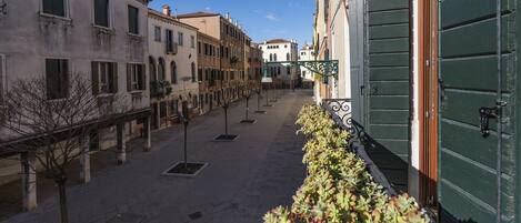 Une rue étroite, bordée de bâtiments des deux côtés, avec une rangée d’arbres et une porte verte sur le côté droit.