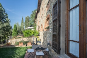 Un bâtiment en pierre avec une porte en bois, un petit patio avec une table et des chaises, et un jardin avec des arbres et des plantes.