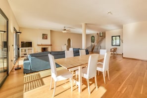A spacious living area with a wooden dining table, white chairs, a blue sofa, and a staircase leading to an upper floor.