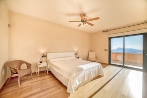 A bedroom with a bed, a chair, a bedside table, a ceiling fan, and a large sliding glass door leading to a balcony with a view.