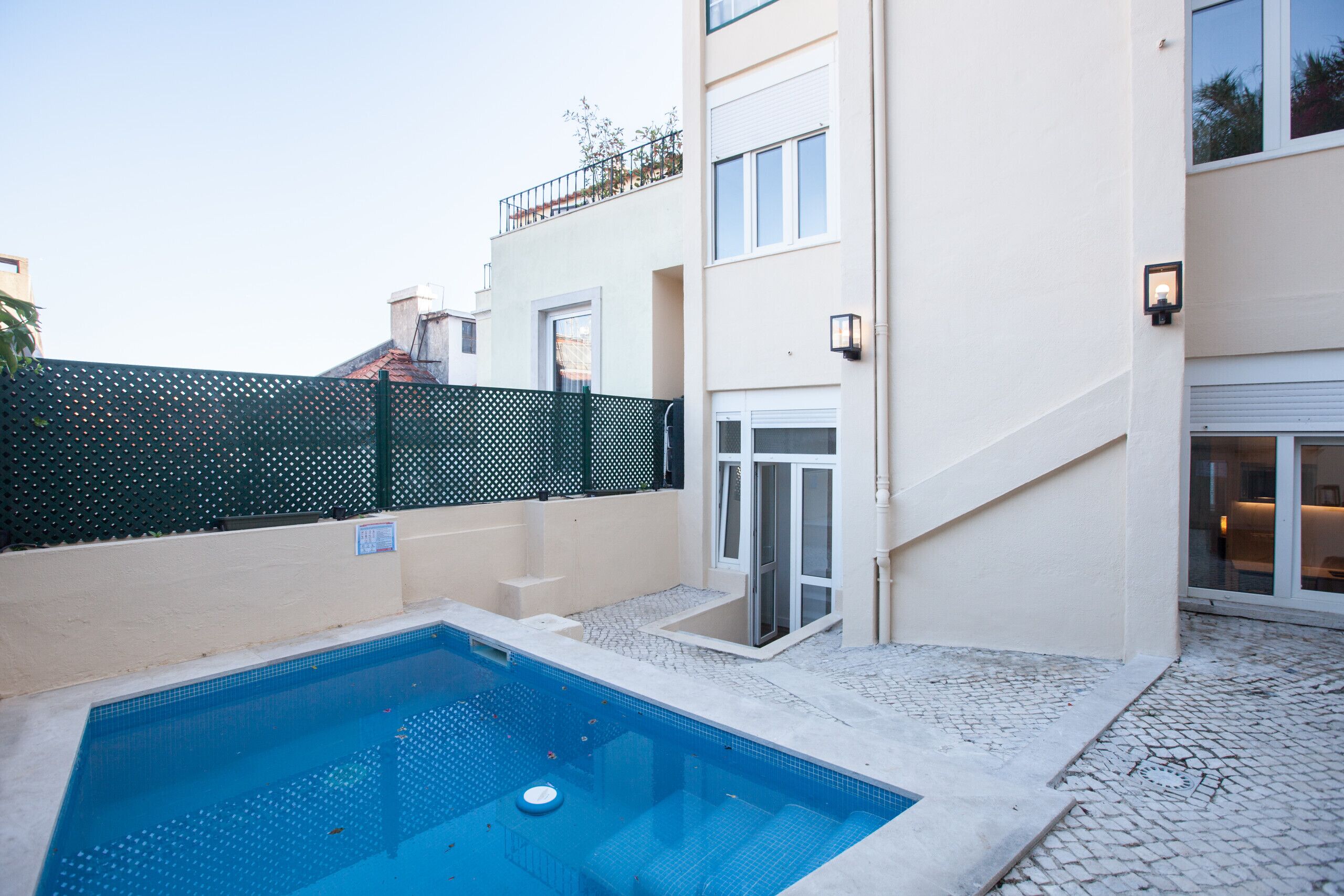 A swimming pool with a tiled deck, a green fence, and a building with a white facade and a glass door.