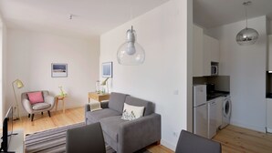 Eine moderne Wohnung mit grauem Sofa, einem Esstisch, einer Küchenzeile inklusive Waschmaschine und einer Pendelleuchte.