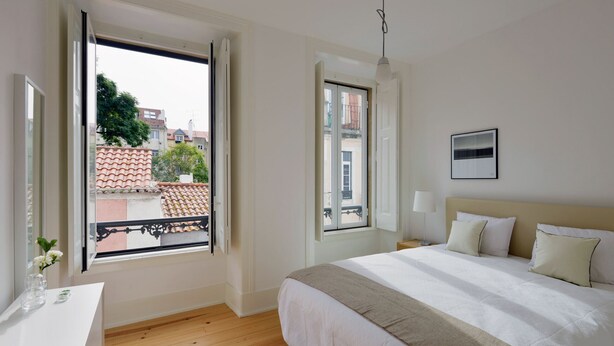 Ein Schlafzimmer mit einem großen Bett, einem Nachttisch, einem Fenster mit Blick auf Gebäude und einer Hängeleuchte.