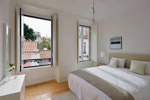 A bedroom with a large bed, a bedside table, a window with a view of buildings, and a hanging light fixture.