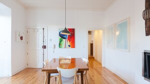 Een eetgedeelte met een houten tafel, witte stoelen, een hanglamp en een kleurrijk abstract schilderij aan de muur.