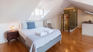 Een slaapkamer met een schuine kap, een bed met een wit en grijs dekbed, een nachtkastje met een lamp en een houten vloer.